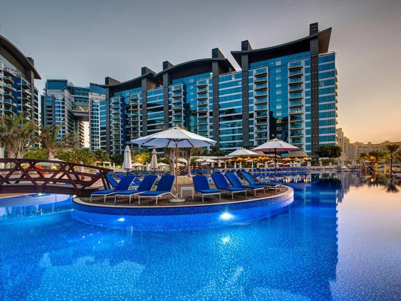 Großer Hotelpool mit blauer Beleuchtung, Liegestühlen und mehrstöckigem Gebäude im Hintergrund.