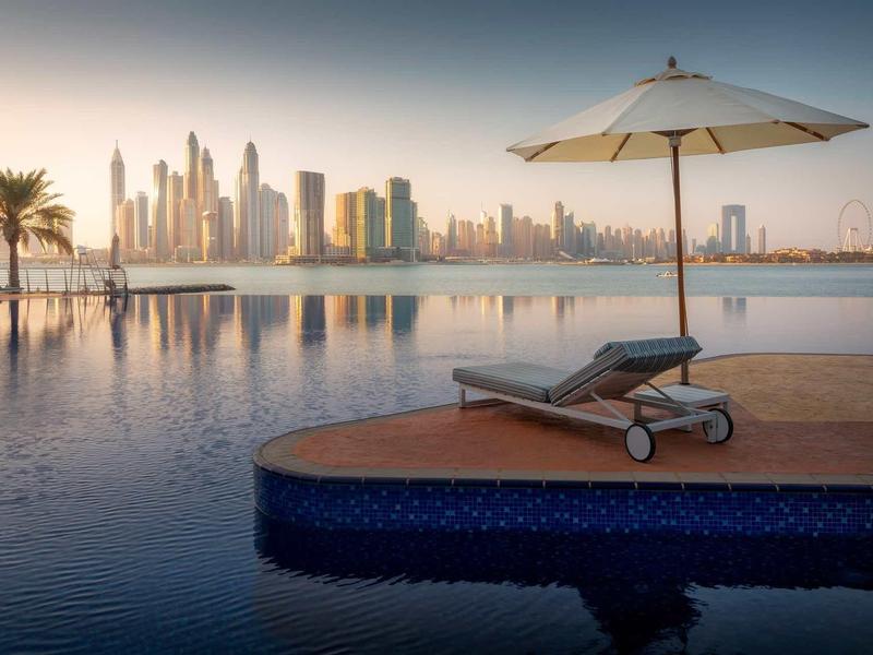 Infinity-Pool mit Liegestuhl und Sonnenschirm, im Hintergrund moderne Hochhäuser bei Sonnenuntergang.