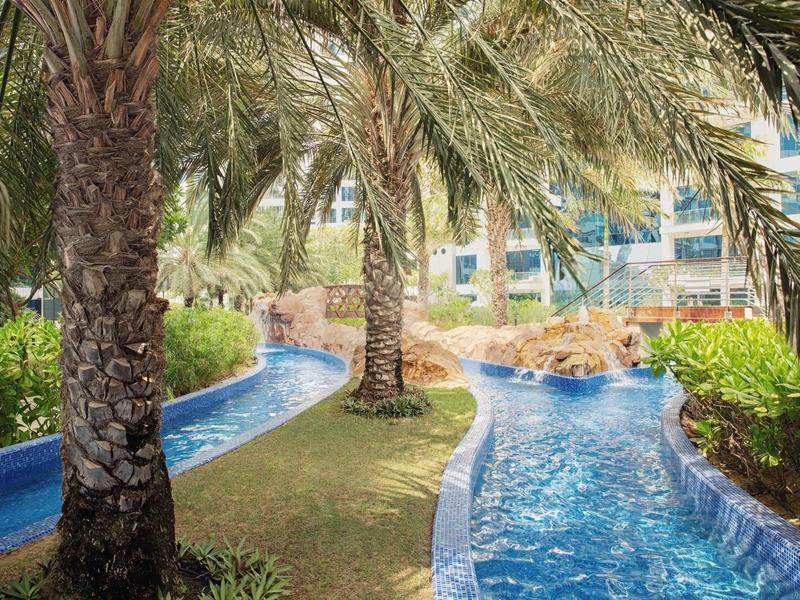 Blauer Wasserkanal schlängelt sich durch grüne Palmen und Büsche unter sonnigem Himmel.