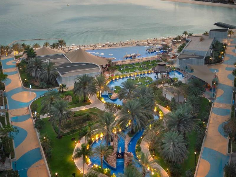 Blick auf einen beleuchteten Resort-Pool mit Palmen und dahinter ruhiges Meer und Stadt am Horizont.