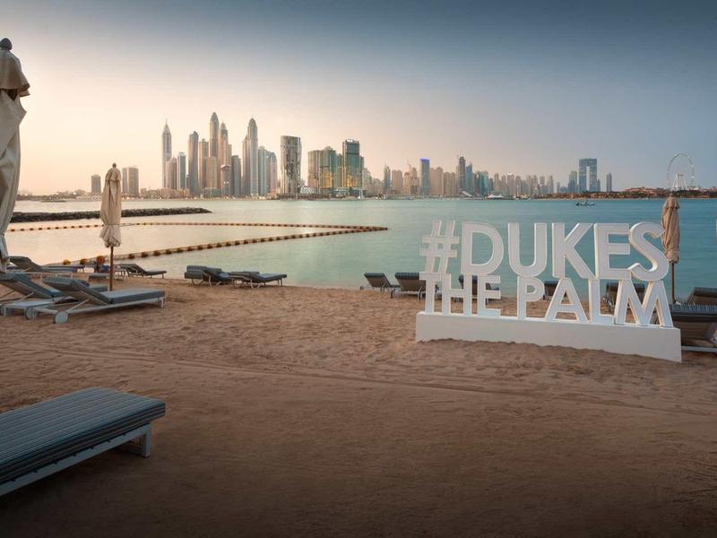 Strand mit Sonnenliegen, geschlossenen Sonnenschirmen und Dubai Skyline im Hintergrund bei Dämmerung.