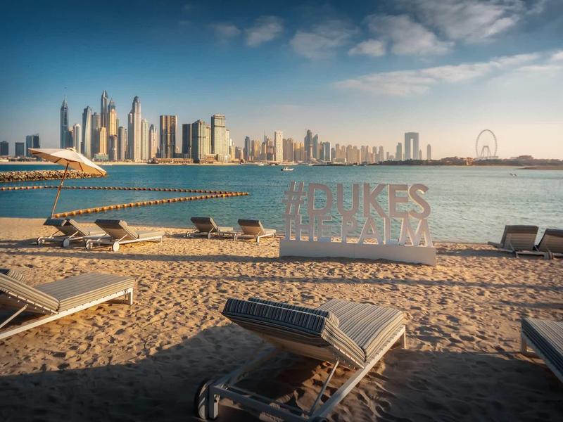 Strand mit Liegen und Blick auf moderne Wolkenkratzer am Wasser bei blauem Himmel.