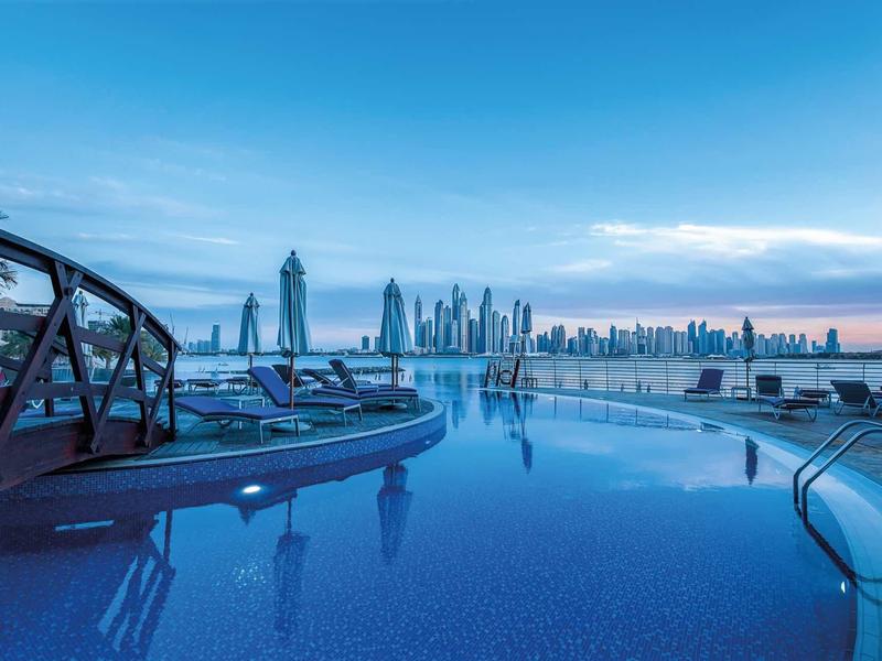 Pool mit klar blauem Wasser, Liegestühle am Rand, moderne Wolkenkratzer im Hintergrund bei Abendhimmel.