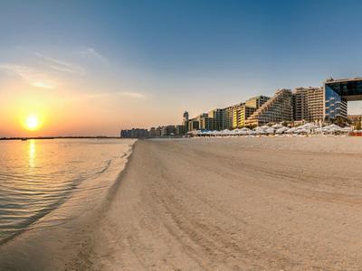 Breiter Sandstrand mit ruhigem Meer und Sonnenuntergang, umgeben von Hotels und Sonnenschirmen.