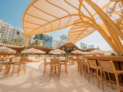 Strandrestaurant mit Holzmöbeln und Sonnenschirmen vor moderner Stadtkulisse.