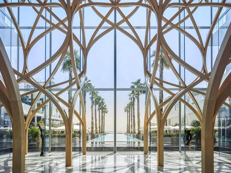 Großer Innenraum mit hohen Holzsäulen und Glasfassade, Blick auf Palmen und Meer.