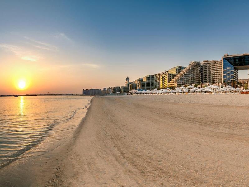 Breiter Sandstrand mit ruhigem Meer und Sonnenuntergang, umgeben von Hotels und Sonnenschirmen.