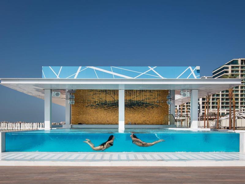 Moderner Pool mit zwei Personen in Liegen vor einer Wasserwand und Hotelgebäuden.