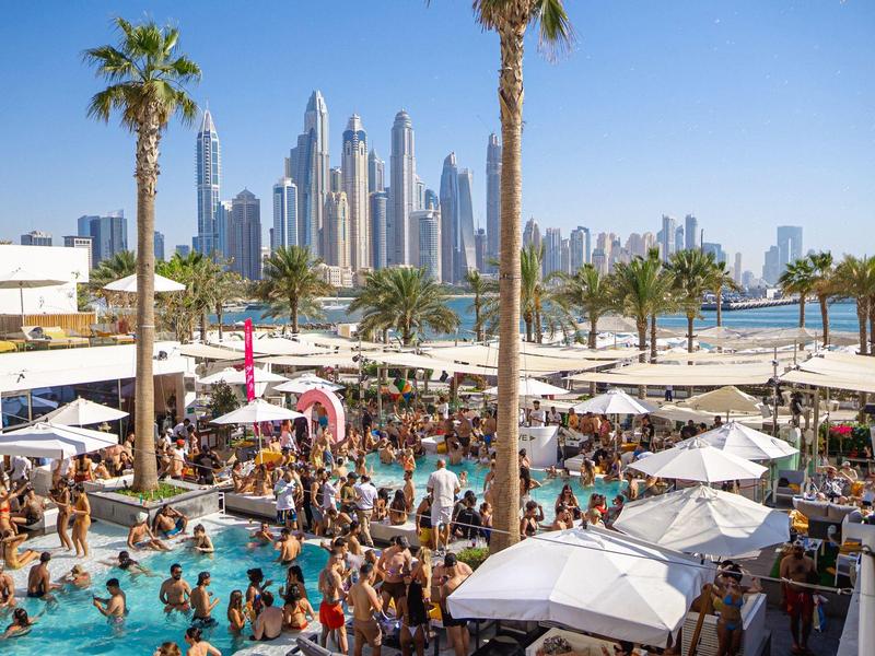 Belebter Poolbereich mit vielen Menschen, Palmen und moderner Stadtsilhouette im Hintergrund.
