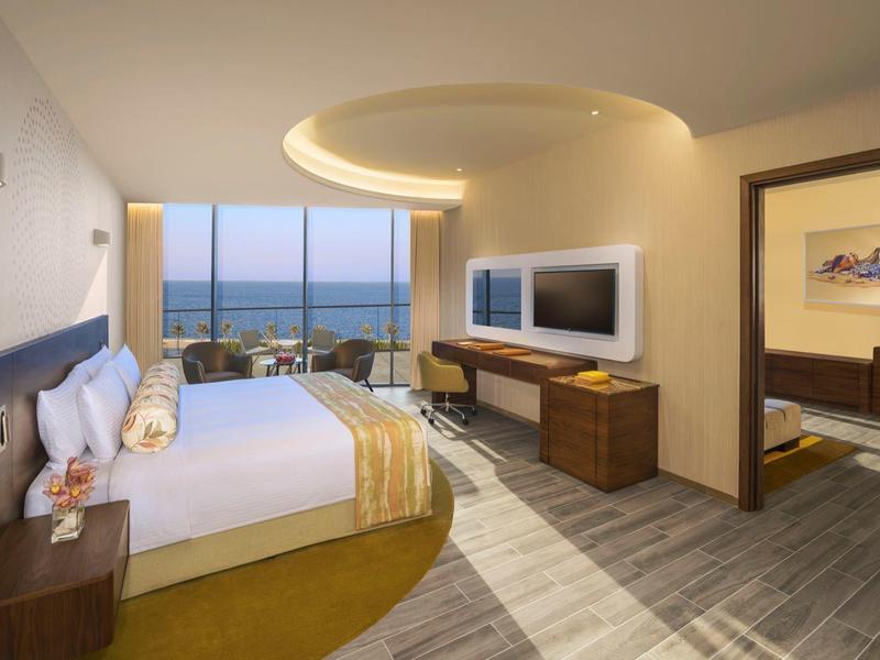 Modernes Hotelzimmer mit großem Bett, Panoramafenster und Blick auf das Meer bei Sonnenuntergang.