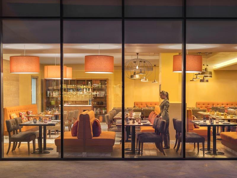 Moderner Gastronomiebereich mit warmem Licht, Tischen, Stühlen und Couch vor großen Glasfenstern.