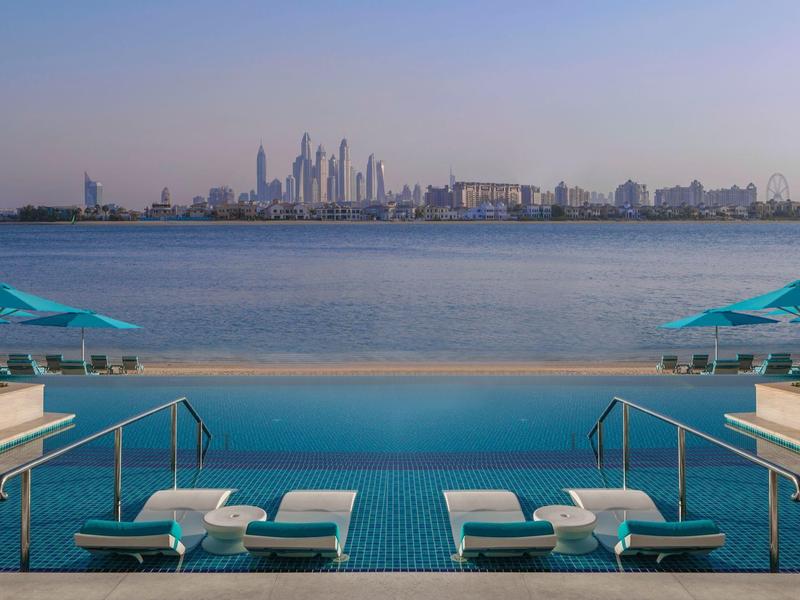 Ein Infinity-Pool mit Liegen und Sonnenschirmen, dahinter ein ruhiges Gewässer und die Skyline einer Stadt.