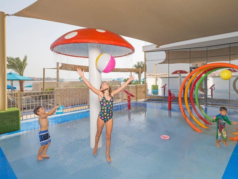 Kinder spielen im flachen Pool mit bunten Wasserspielen und Pilz-Wassersprudler.