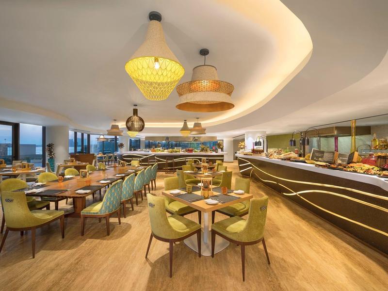 Elegantes Restaurant mit runden Tischen, grünen Sesseln, modernem Buffetbereich und warmem Licht.