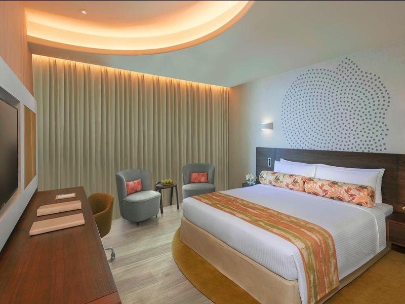 Modernes Schlafzimmer mit Doppelbett, Sessel, Wand-TV und weichen Vorhängen in warmem Licht.