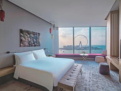 Modernes Hotelzimmer mit King-Size-Bett, großem Fenster und Blick auf ein Riesenrad bei Sonnenuntergang.