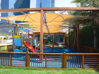 Kinderspielplatz mit buntem Klettergerüst, orangenen Sonnensegeln und grüner Wiese im Vordergrund.