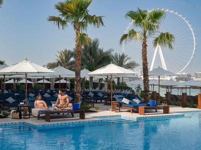 Poolanlage mit blauen Liegen, Palmen, Sonnenschirmen und großem Riesenrad im Hintergrund.