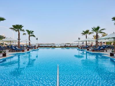 Großer Pool mit blauen Fliesen, umgeben von Liegen und Palmen unter klarem Himmel.
