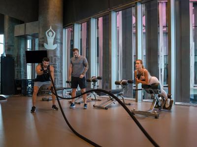 Drei Personen trainieren in hellem Fitnessstudio mit großen Fenstern und Kampfbändern.