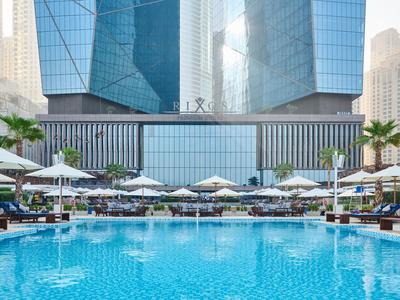 Großer blauer Pool mit weißen Sonnenschirmen und Liegen vor modernen Glasgebäuden in der Stadt.