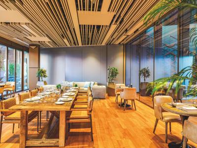 Modern restaurant with wooden floors, long tables, and large windows overlooking greenery.