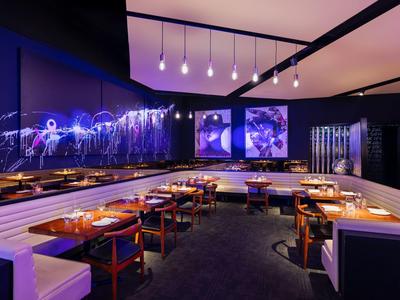 Restaurante moderno con iluminación violeta, mesas de madera y bancos blancos en un hotel.