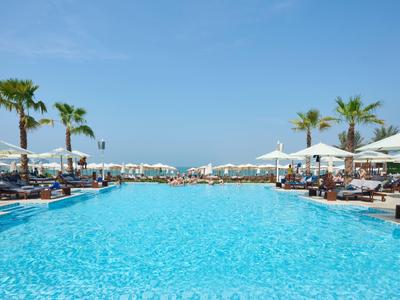 Großer Hotelpool mit Liegestühlen und Palmen unter blauem Himmel.