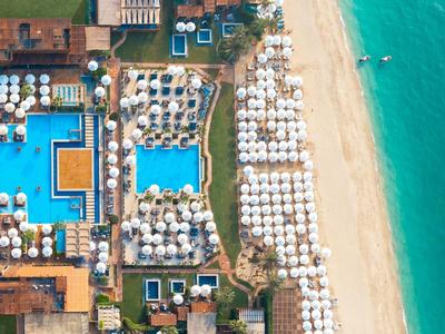 Vogelperspektive eines Resorts mit Pool, Sonnenschirmen und Strand am türkisfarbenen Meer.