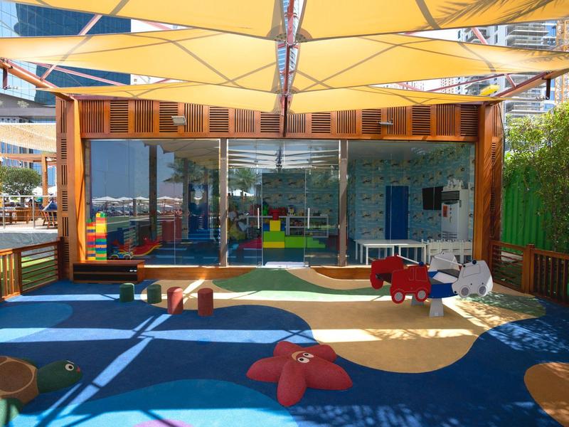 Área de juegos al aire libre con suelo azul y amarillo, juguetes y estructura de madera para niños en hotel.