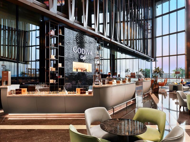 Área de cafetería moderna de un hotel con grandes ventanales y bar elegante en tonos cálidos.