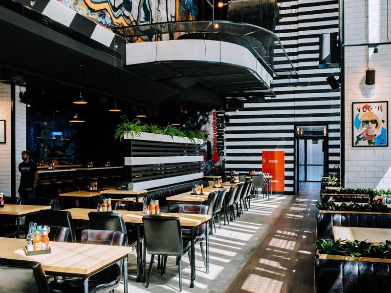 Interior moderno de restaurante con sillas negras, mesas de madera y diseño geométrico en la pared.