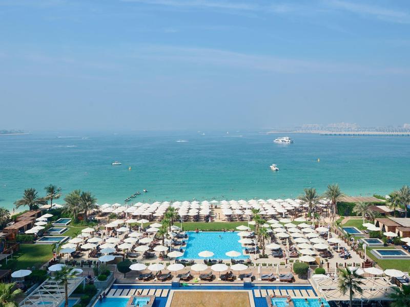 Vista de una piscina de hotel con sombrillas y el mar al fondo bajo un cielo despejado.