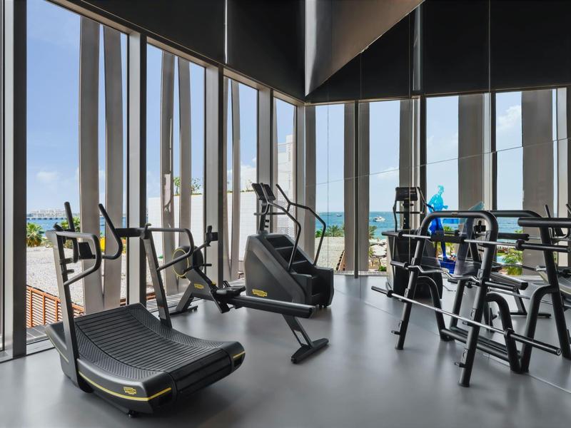 Equipos modernos de gimnasio en una sala de fitness de hotel con ventanas de piso a techo y vista al mar.