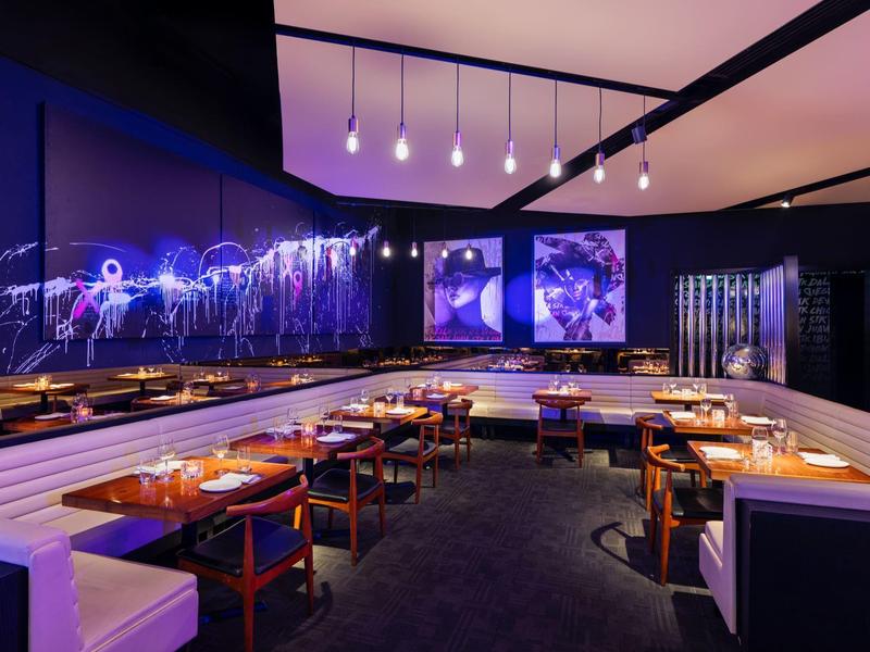 Restaurante moderno con iluminación violeta, mesas de madera y bancos blancos en un hotel.