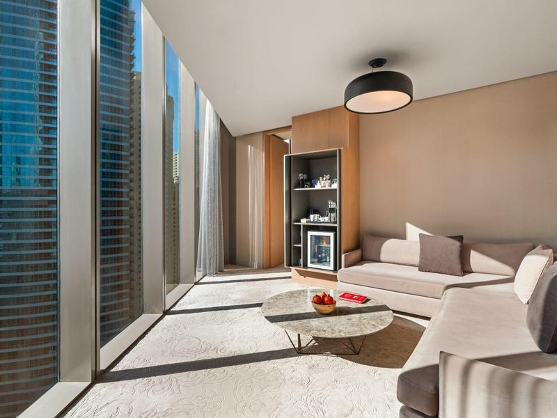 Salón de hotel luminoso con ventanas de piso a techo, sofá claro y mesa de centro moderna con aperitivos.