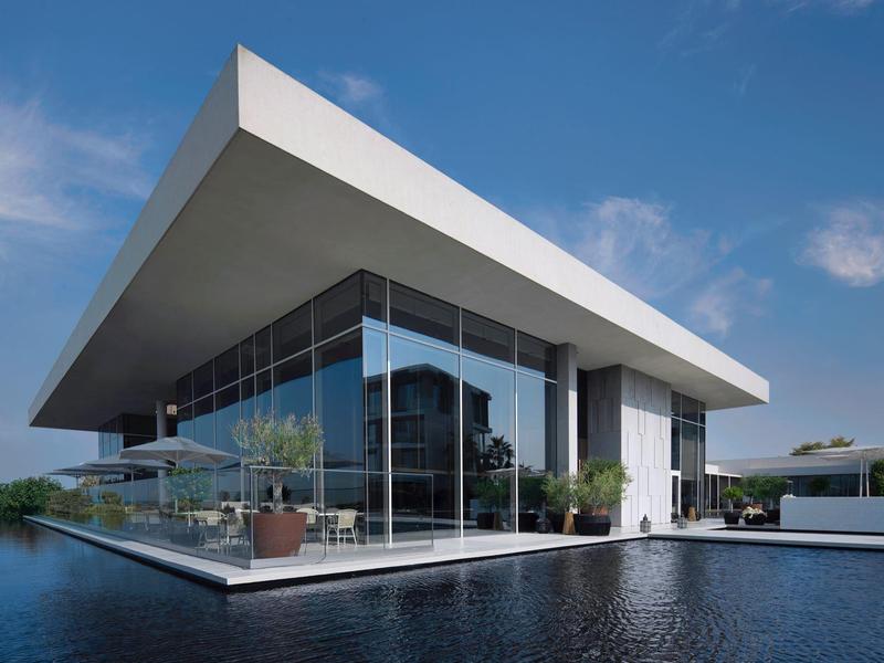 Modernes Gebäude mit schrägem Dach über Wasser, umgeben von Glasfronten und Pflanzen.