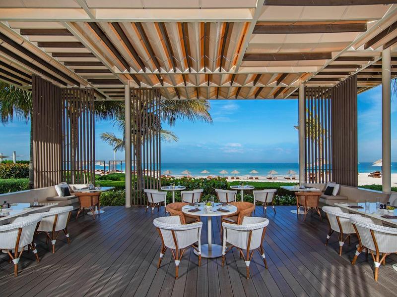 Offener, moderner Restaurantbereich mit Holzboden, weißen Stühlen und Blick auf Meer und Strand.