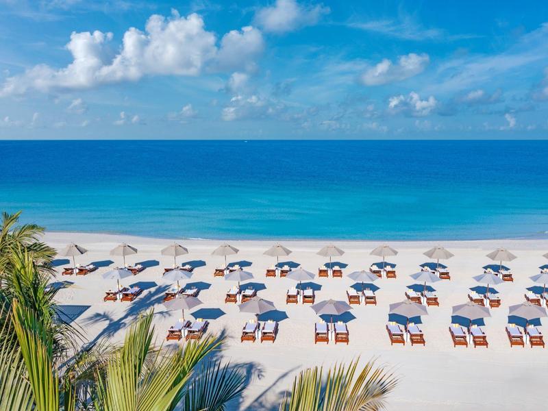 Weißer Sandstrand mit Sonnenliegen und Sonnenschirmen vor klarem blauem Meer und Himmel.