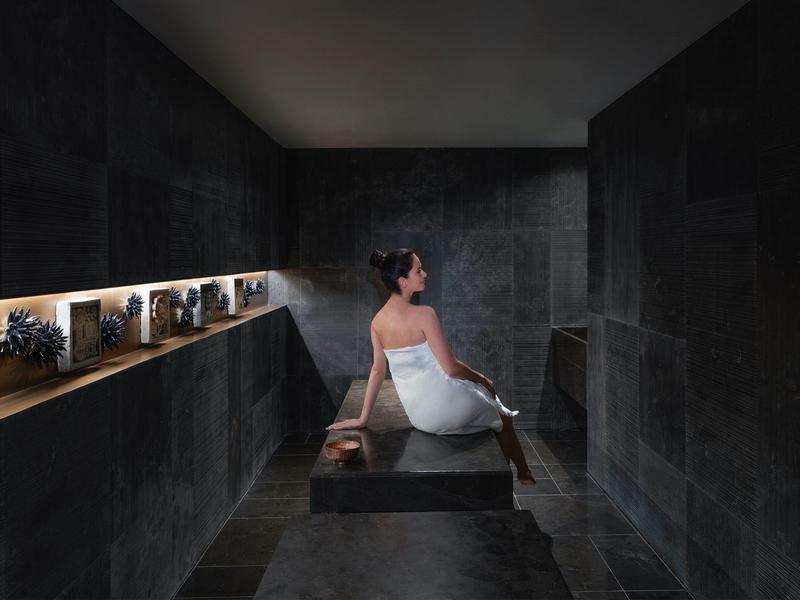 Eine Frau in einem weißen Handtuch sitzt in einem dunklen Dampfbad mit schwarzen Wänden und indirekter Beleuchtung.