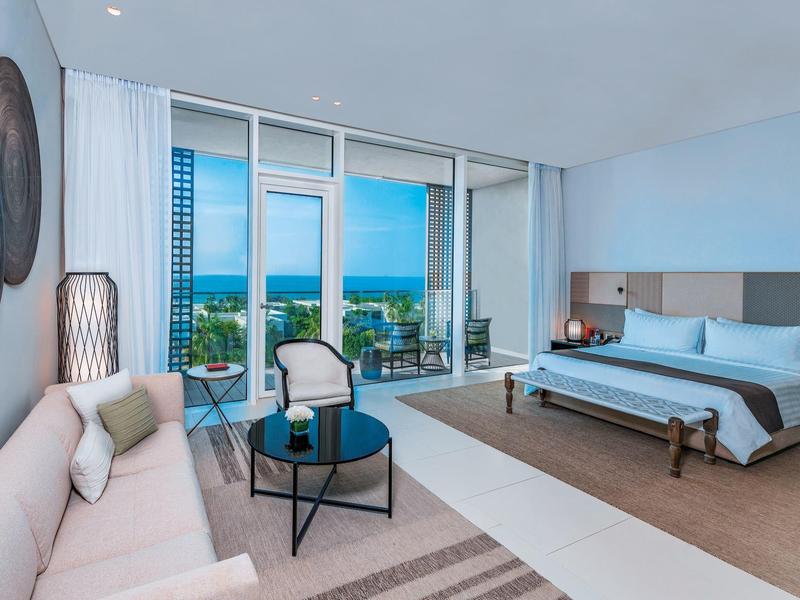 Helles Hotelzimmer mit großem Bett, Sofa, Tisch und Meerblick durch große Fenster.