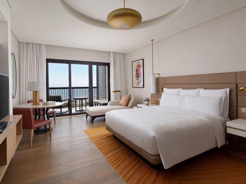 Helles Hotelzimmer mit großem Doppelbett, Fernseher, Schreibtisch, Sofa und Balkon mit Blick.