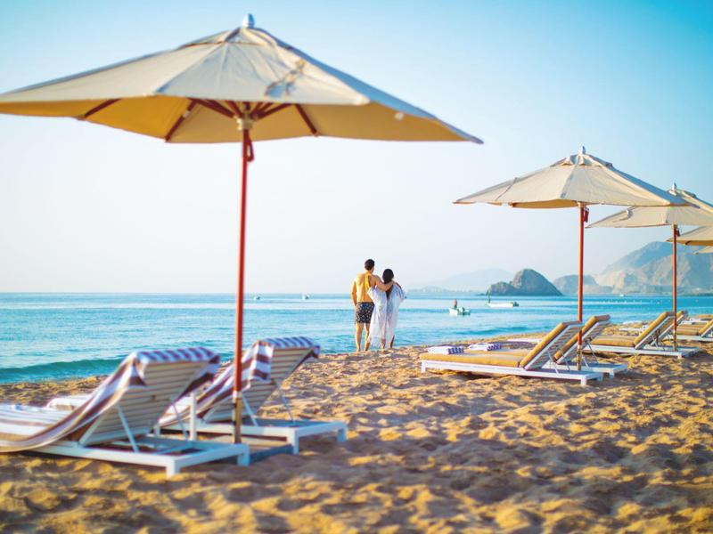 Strand mit Sand, Liegen und Sonnenschirmen, Paar geht am Wasser entlang, blauer Himmel.