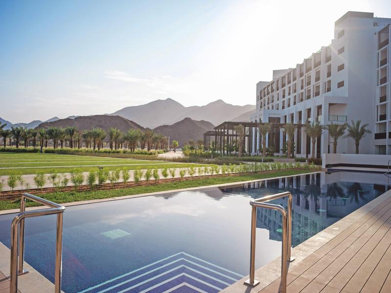 Moderner Hotelkomplex mit großem Pool, Palmen und Bergkulisse im Hintergrund bei Sonnenschein.