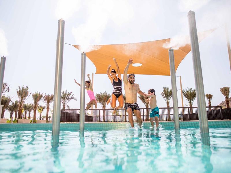 Zwei Erwachsene und ein Kind springen lachend von einem Beckenrand ins Wasser unter blauem Himmel.