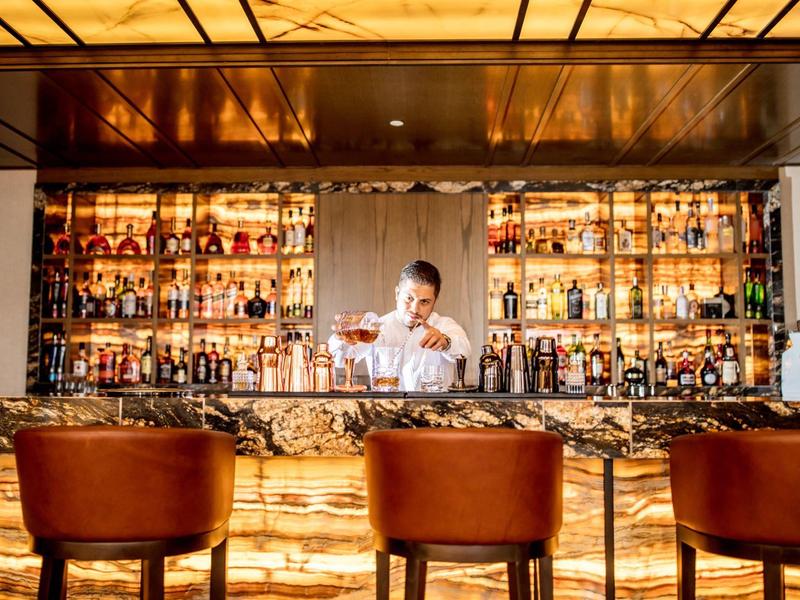 Bartender mixt Getränke an beleuchtetem Bar-Tresen mit drei Lederhockern im Vordergrund.