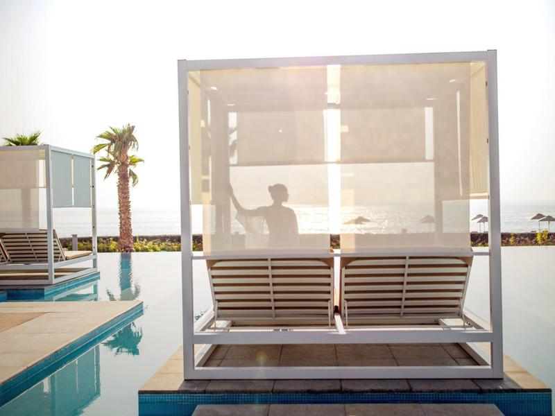 Moderner, überdachter Strandliegenplatz mit Holzbank und Glaswänden neben einem Pool bei Sonnenuntergang.