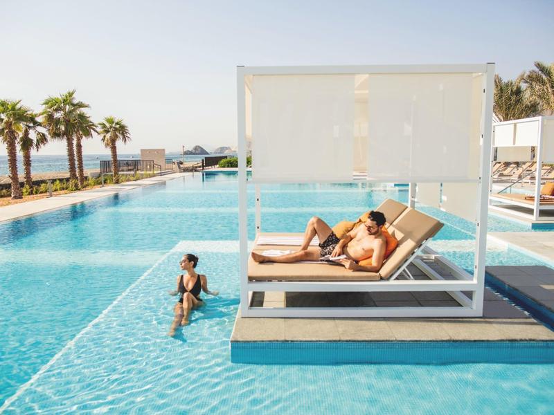 Ein Mann entspannt auf einer Liege in einem modernen Poolbereich mit Palmen und Meer im Hintergrund.