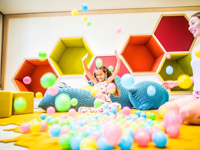 Kind spielt mit bunten Bällen in hellem Raum mit gelbem Boden und sechseckigen Wandregalen.
