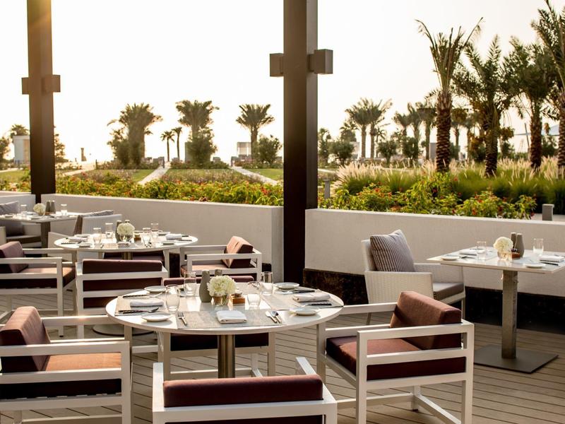 Terrassenrestaurant mit runden Tischen, Stühlen mit Kissen, Palmen und Sonnenlicht im Hintergrund.